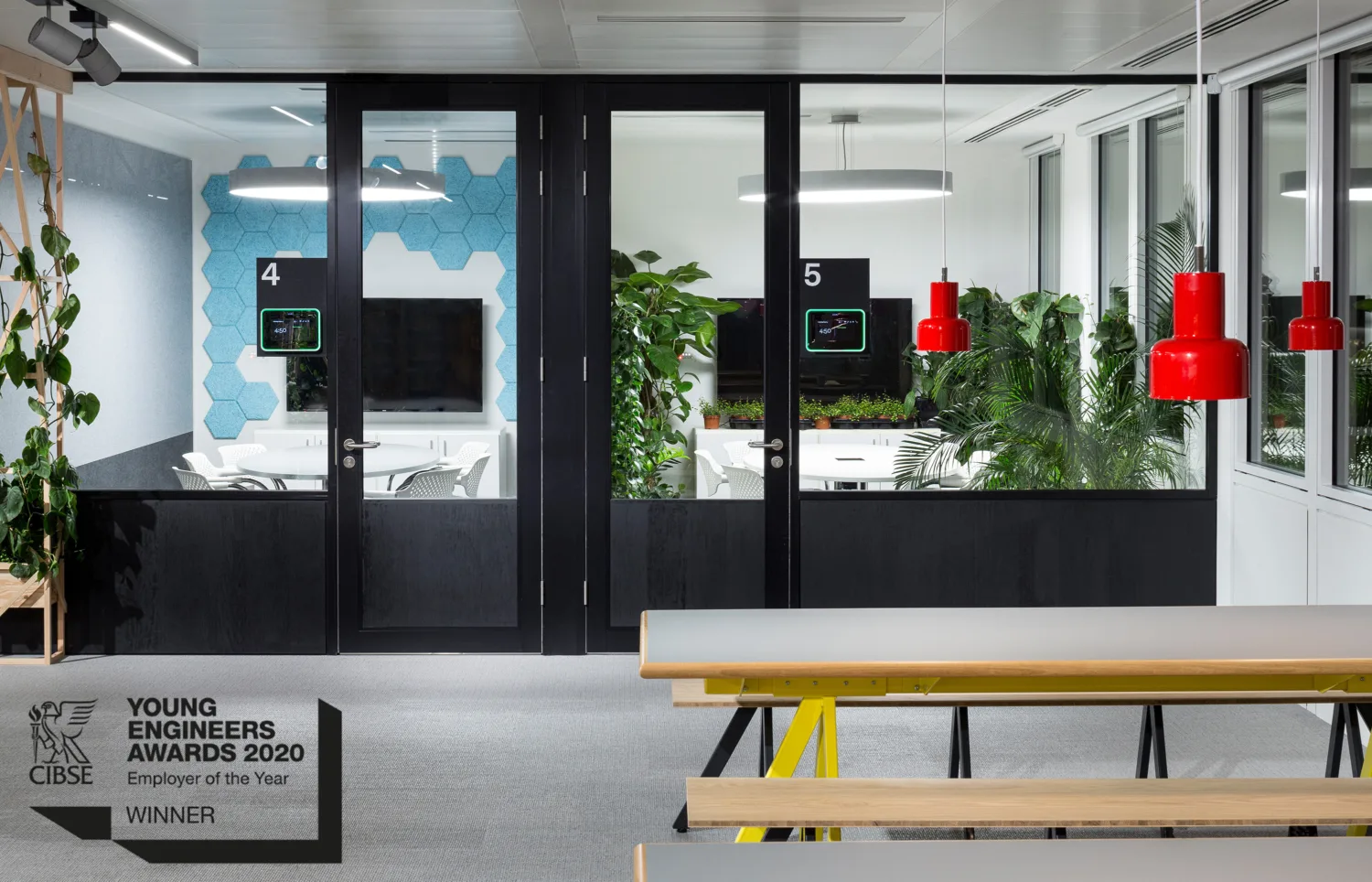 Interior of Cundall London office featuring two meeting rooms with black metallic doors, in front of which two grey and wooden dining tables are visible underneath red modern hanging lighting fixtures, in the bottom left corner is the CIBSE logo