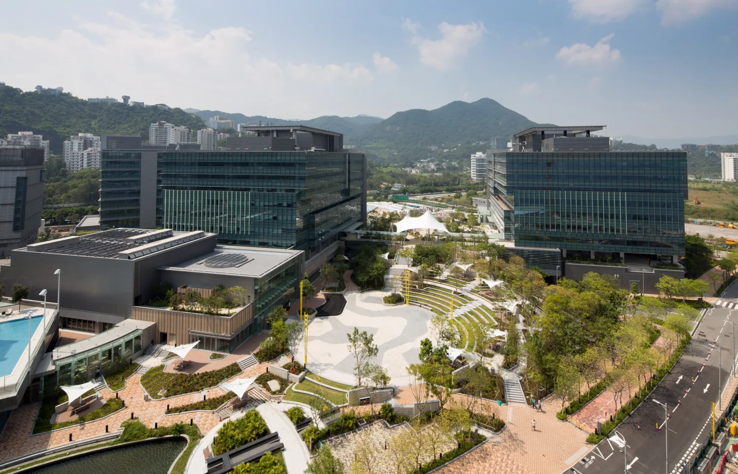 A collection of large modern buildings in a park of cream pathways and vegetation, in the background are green hills and mountains