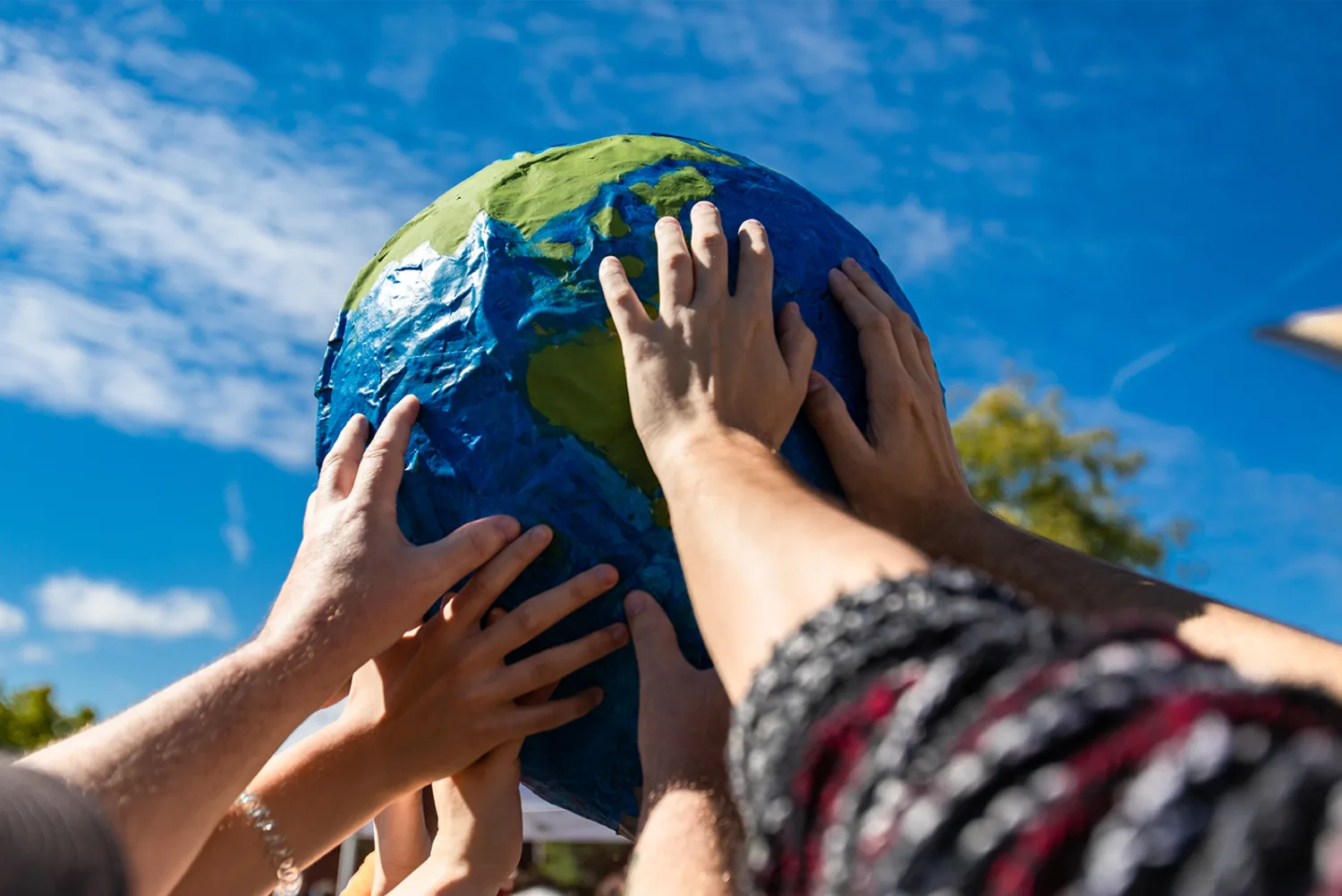 View from below of a painted textured globe, supported by multiple people with their hands outstretched, against a blue slightly cloudy sky