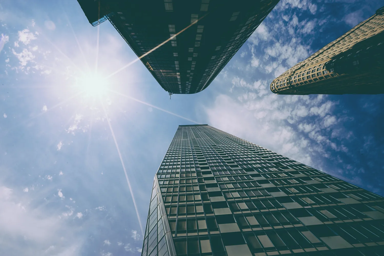 View from below of three tall buildings against a blue slightly cloudy sky and a sun that is forming a 'lens flare' effect
