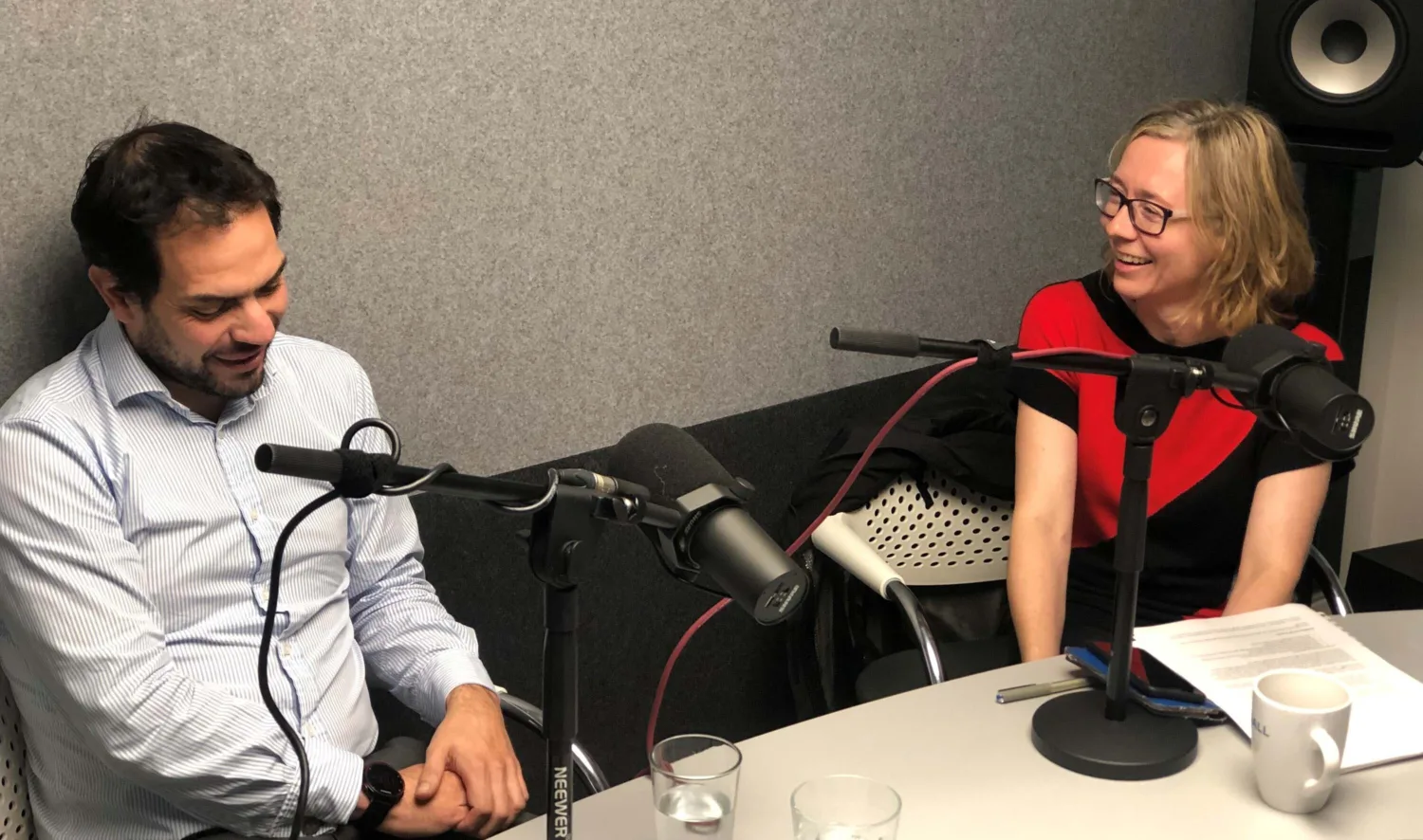 Two people sat at a table behind desktop microphones with scripts, mugs and glasses of water