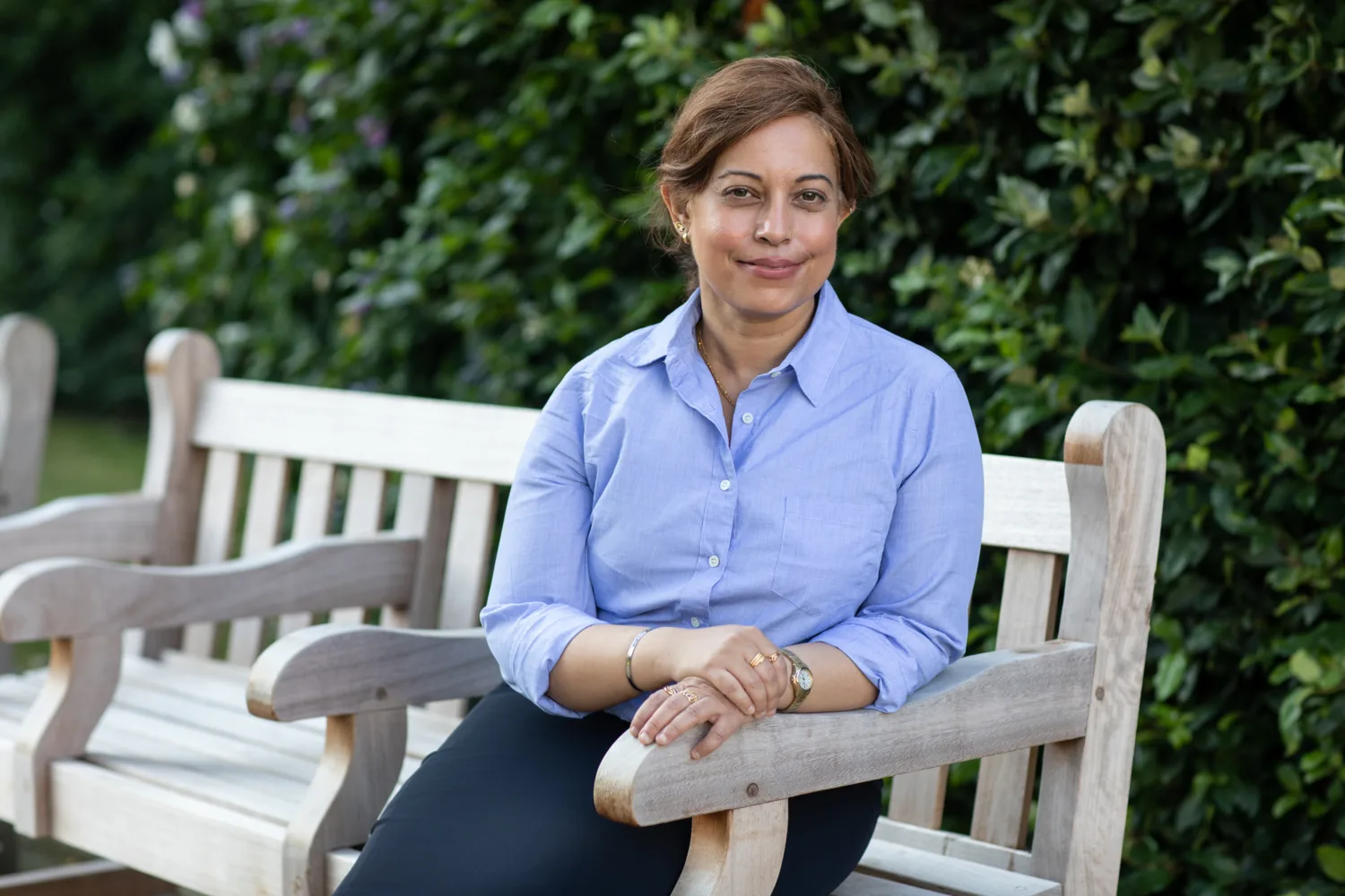 Kavita wearing light blue shirt sat on a wooden bench in front of a tall green hedge