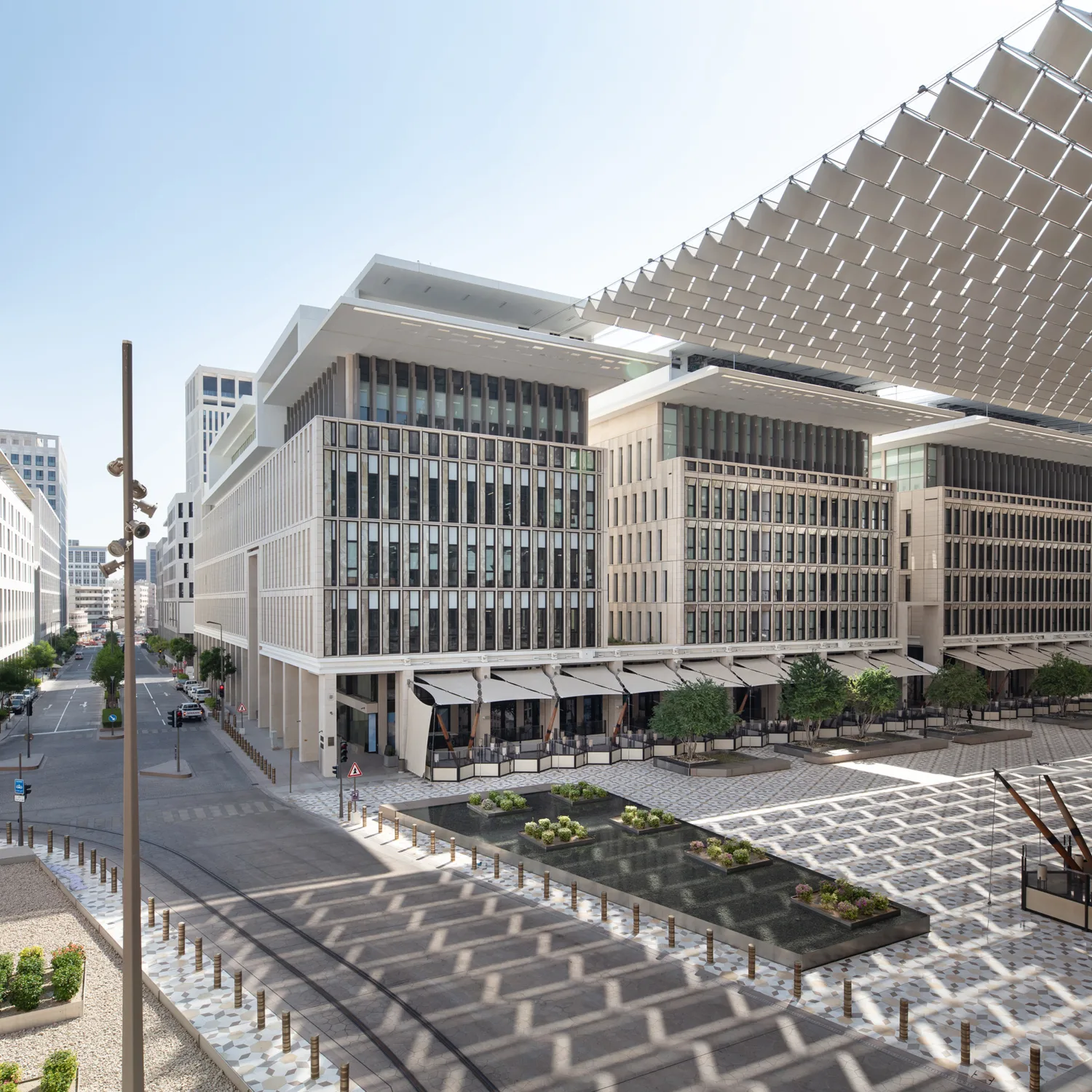 Modern urban plaza with mid-rise buildings featuring glass windows and white facades, patterned pavement, landscaped plant areas, and a large overhead shade structure.
