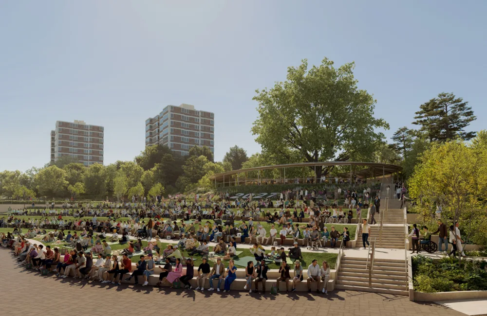 Outdoor amphitheater with people seated on tiered grassy steps; central staircase leads to covered area; three tall apartment buildings and trees in background under clear blue sky.