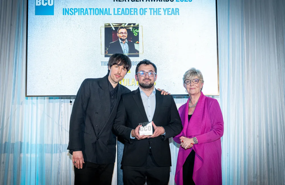 Alt text: Three people standing in front of a large screen displaying “BCO NextGen Awards 2025 Inspirational Leader of the Year” with a photo and text underneath. The person in the center is holding a rectangular award with the letters “BCO” visible. The background includes white draped curtains and bright lighting.