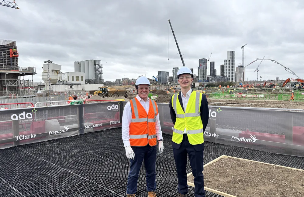Alec Stewart stood on the left and Tom Heywood on right wearing hard hats, high‑visibility vests, long‑sleeved shirts, trousers, and work boots stand on a raised platform overlooking an active construction site with cranes, machinery, and high‑rise buildings in the background.