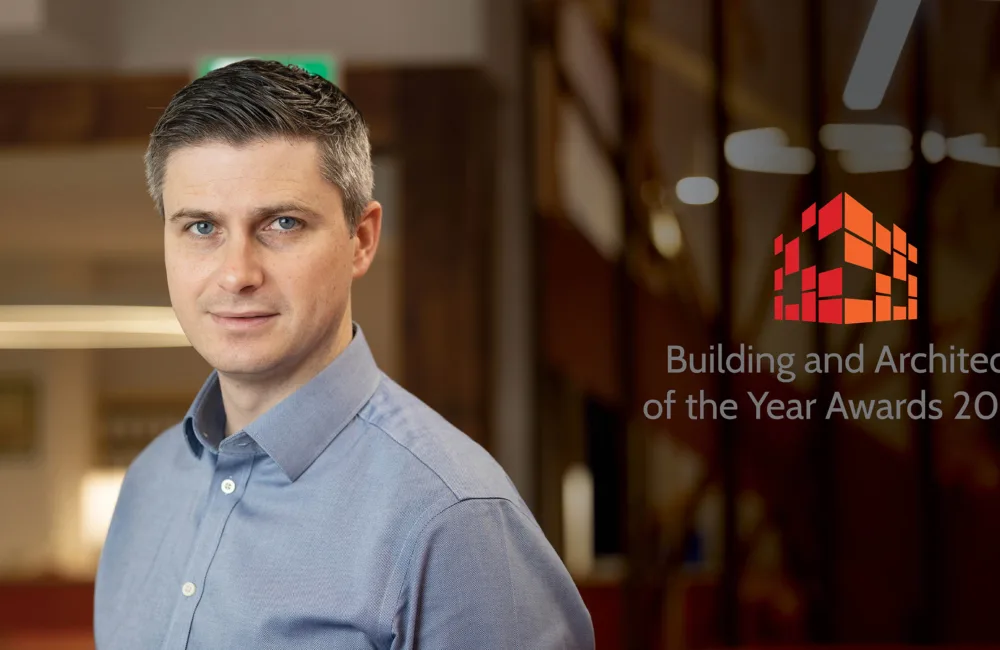 A person wearing a light blue collared shirt stands indoors beside a glass wall displaying a red geometric logo and the text “Building and Architect of the Year Awards 2026,” with warm lighting and wooden details in the background.