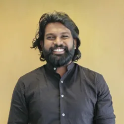 Jotham stands against a plain light‑wood background wearing a dark long‑sleeved shirt with the sleeves rolled up.