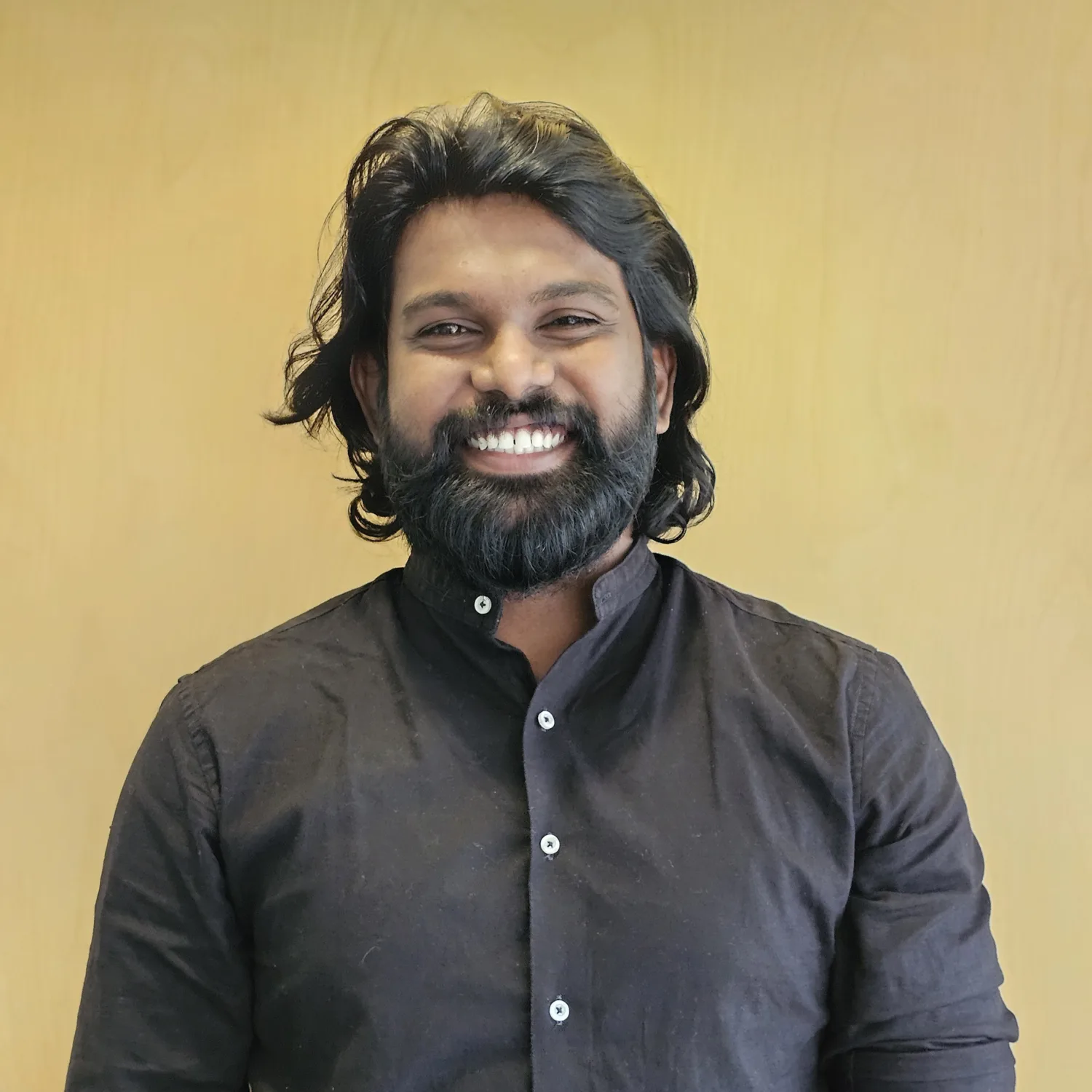 Jotham stands against a plain light‑wood background wearing a dark long‑sleeved shirt with the sleeves rolled up.