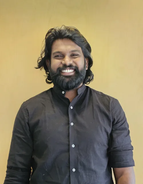 Jotham stands against a plain light‑wood background wearing a dark long‑sleeved shirt with the sleeves rolled up.
