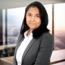 A cut out of Priya in a white shirt and grey suit jacket superimposed on an AI office background