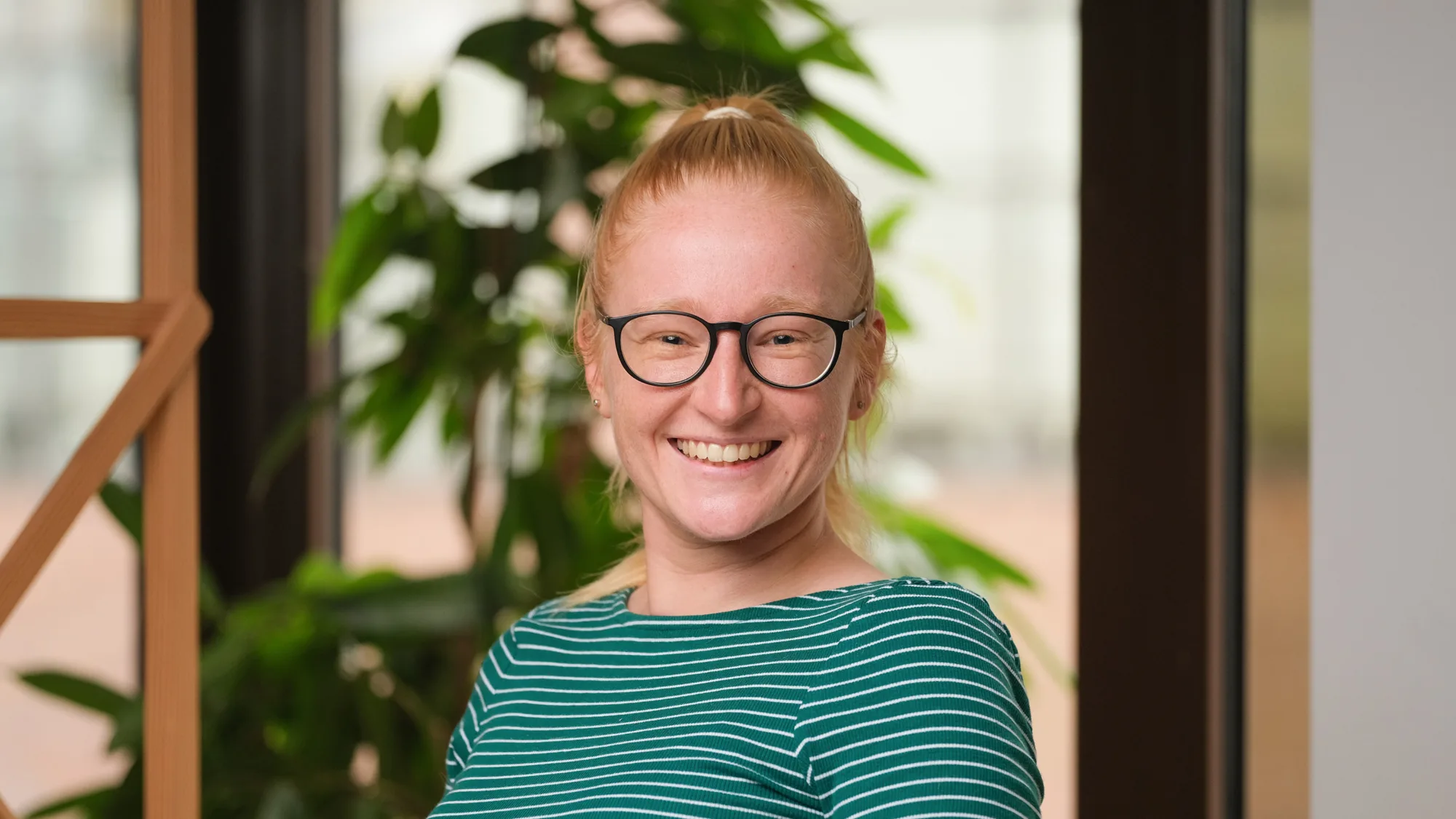 Alison smiling to camera in a green striped long sleeve top in front of a wooden trellis and plant