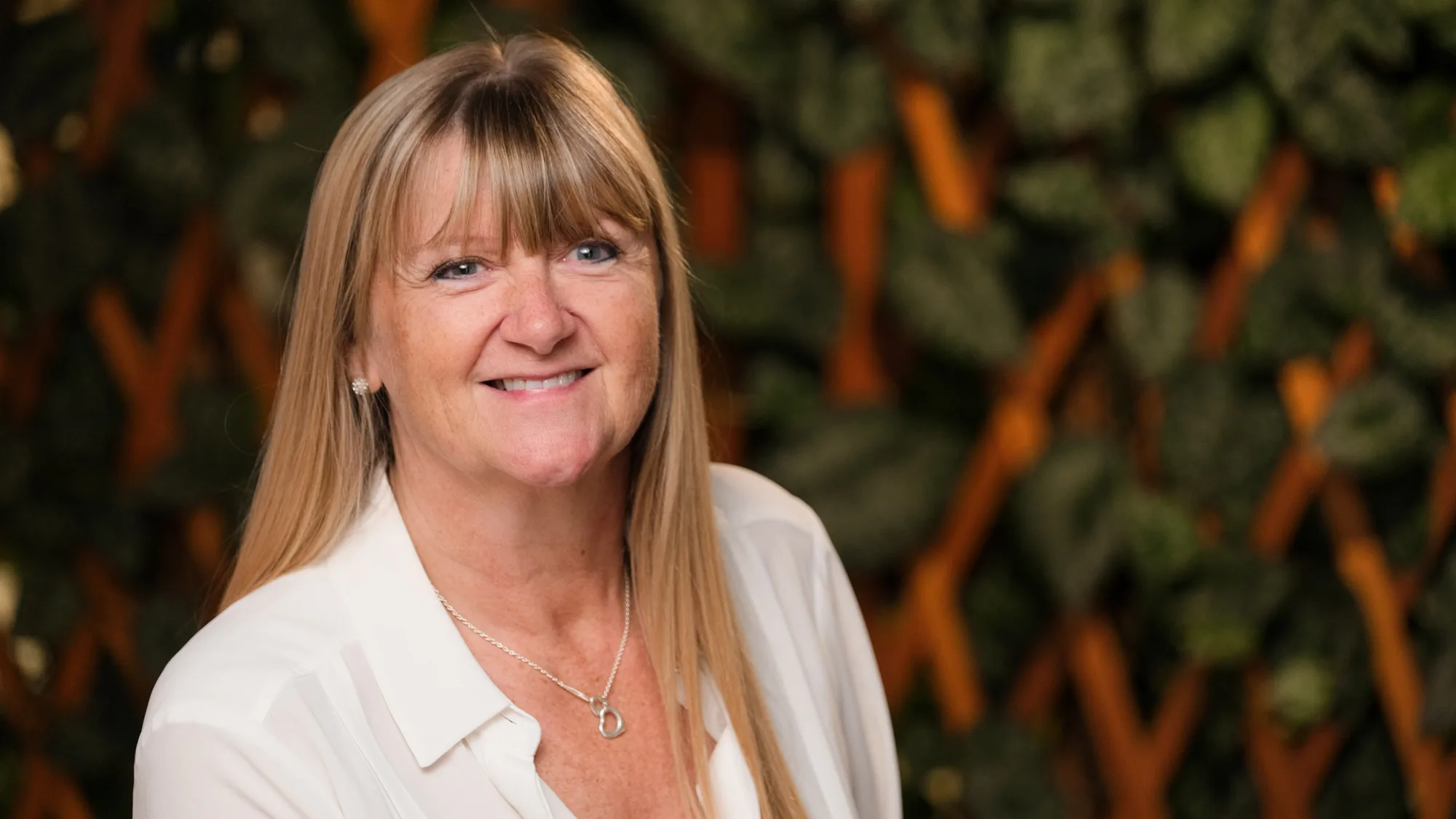 Belinda in a white blouse smiling to camera in front of a trelis wall