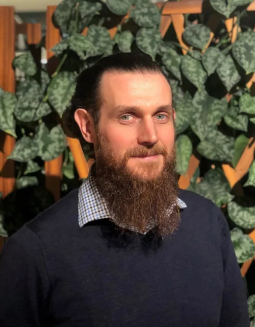 Indoor portrait of a person with long hair tied back and a full beard, wearing a dark sweater over a checkered collar. The background features a wooden lattice partially covered with green leafy plants.