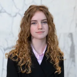 Portrait photograph of a person with long, curly ginger hair wearing a black jacket over a light pink collared shirt, standing against a pale grey and white marble wall background.