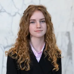 Portrait photograph of a person with long, curly ginger hair wearing a black jacket over a light pink collared shirt, standing against a pale grey and white marble wall background.
