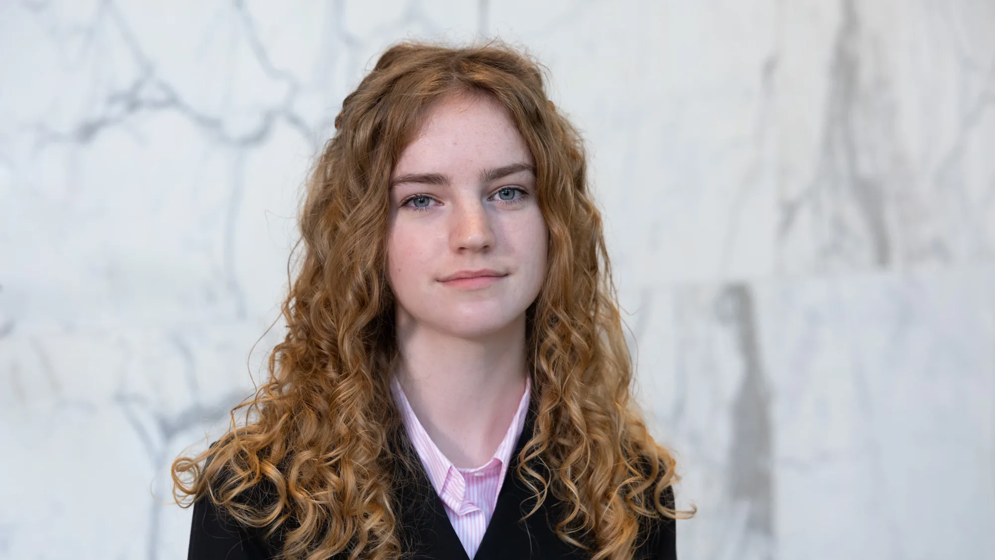 Portrait photograph of a person with long, curly ginger hair wearing a black jacket over a light pink collared shirt, standing against a pale grey and white marble wall background.