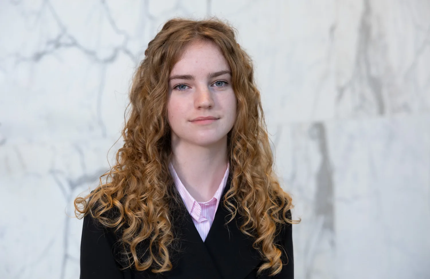 Portrait photograph of a person with long, curly ginger hair wearing a black jacket over a light pink collared shirt, standing against a pale grey and white marble wall background.