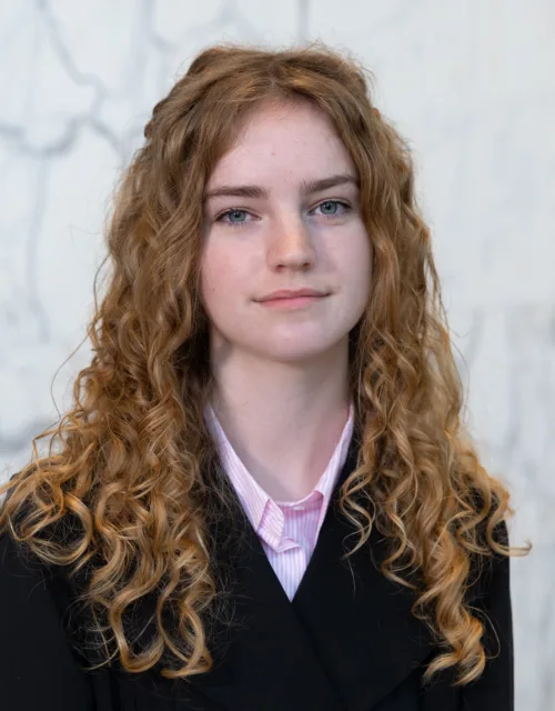 Portrait photograph of a person with long, curly ginger hair wearing a black jacket over a light pink collared shirt, standing against a pale grey and white marble wall background.
