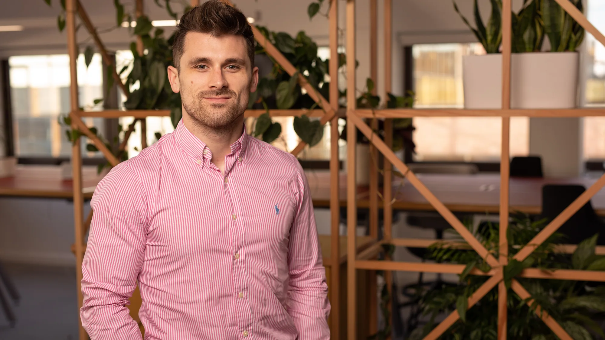 Ian stood with hands in pockets in a pink long sleeve shirt in front of a plant trellis in an office