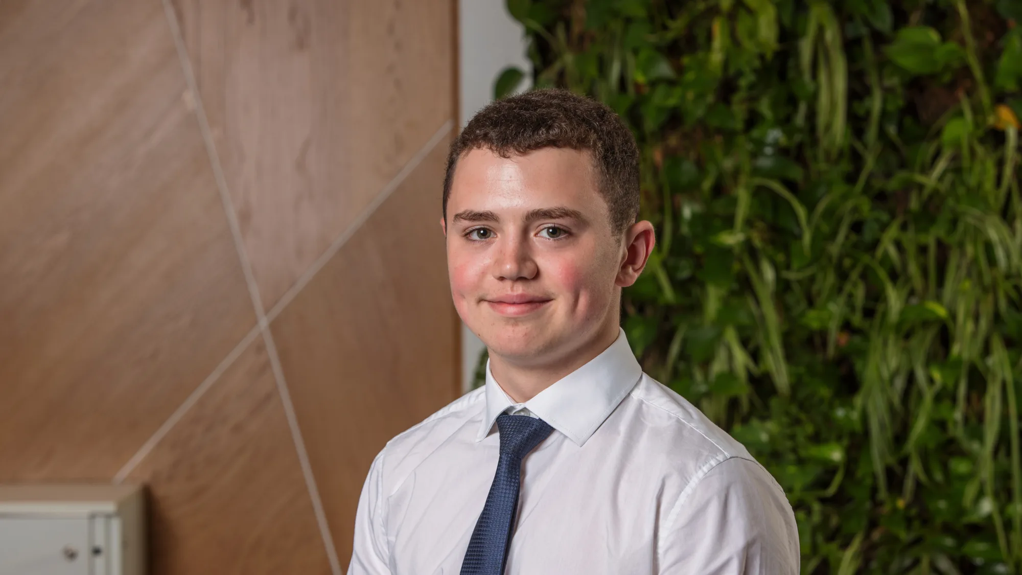 Person wearing a white dress shirt and dark patterned tie is seated indoors. Background features a wooden panel with diagonal lines on the left and a green vertical plant wall on the right.