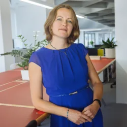 A person in a blue dress stands beside a large shared workspace table in a modern, open‑plan office with plants, shelving, and desks visible in the background.