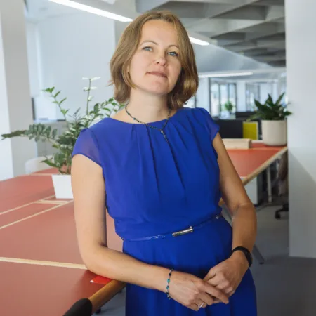 A person in a blue dress stands beside a large shared workspace table in a modern, open‑plan office with plants, shelving, and desks visible in the background.
