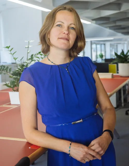 A person in a blue dress stands beside a large shared workspace table in a modern, open‑plan office with plants, shelving, and desks visible in the background.