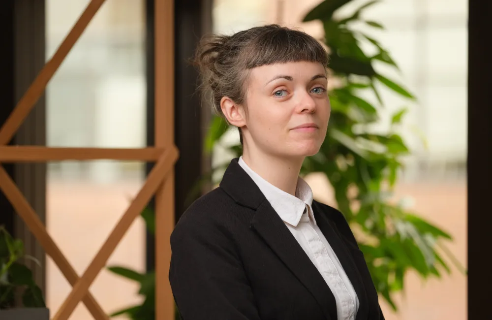 Kim standing in front of wooden trellis wearing a black blazer and a white shirt.