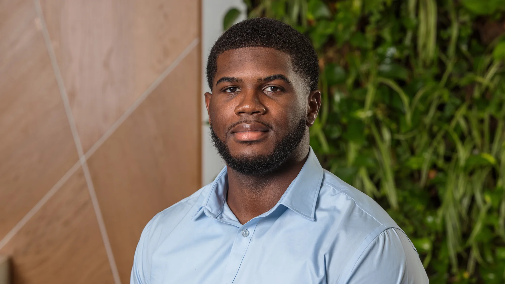 Portrait of a person wearing a light blue button-up shirt, facing the camera indoors. Background shows a wooden panel with diagonal lines and a wall of green plants.