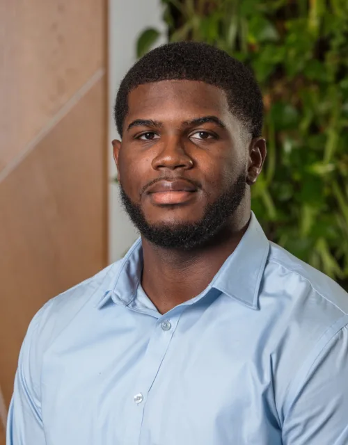 Portrait of a person wearing a light blue button-up shirt, facing the camera indoors. Background shows a wooden panel with diagonal lines and a wall of green plants.