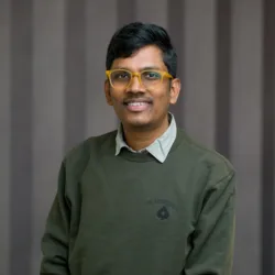 Person wearing a dark green crew-neck sweatshirt with a small emblem on the left chest and a light grey collared shirt underneath, seated against a background of vertical dark grey panels.