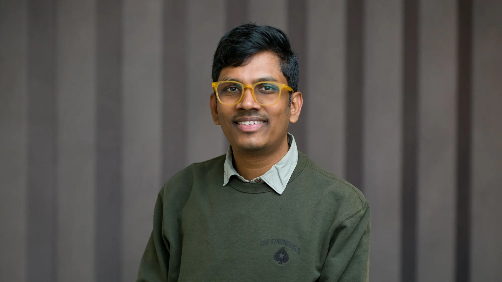 Person wearing a dark green crew-neck sweatshirt with a small emblem on the left chest and a light grey collared shirt underneath, seated against a background of vertical dark grey panels.