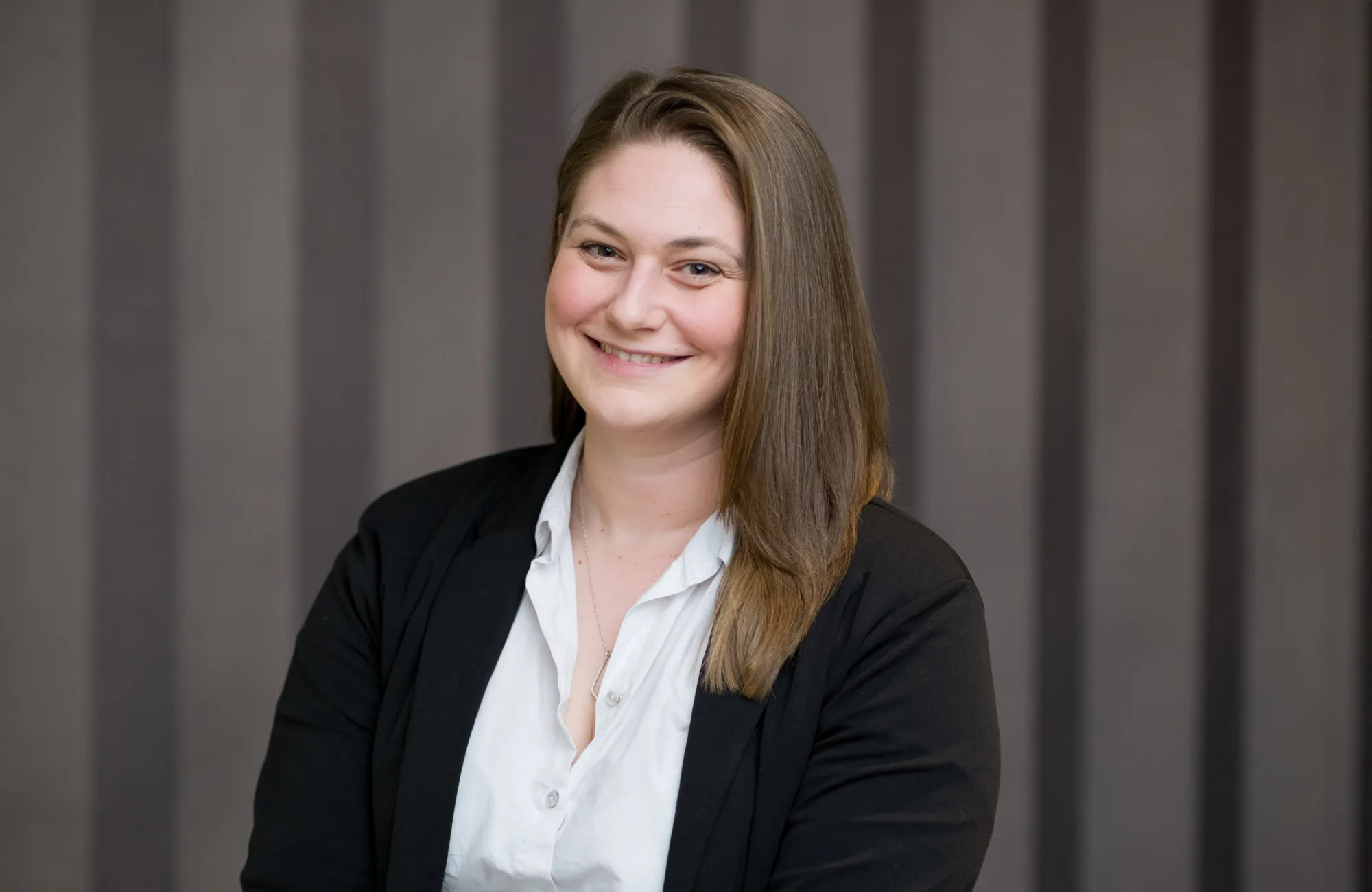 A person with shoulder-length brown hair, wearing a white button-up shirt and black blazer, seated in front of a background with vertical grey stripes.