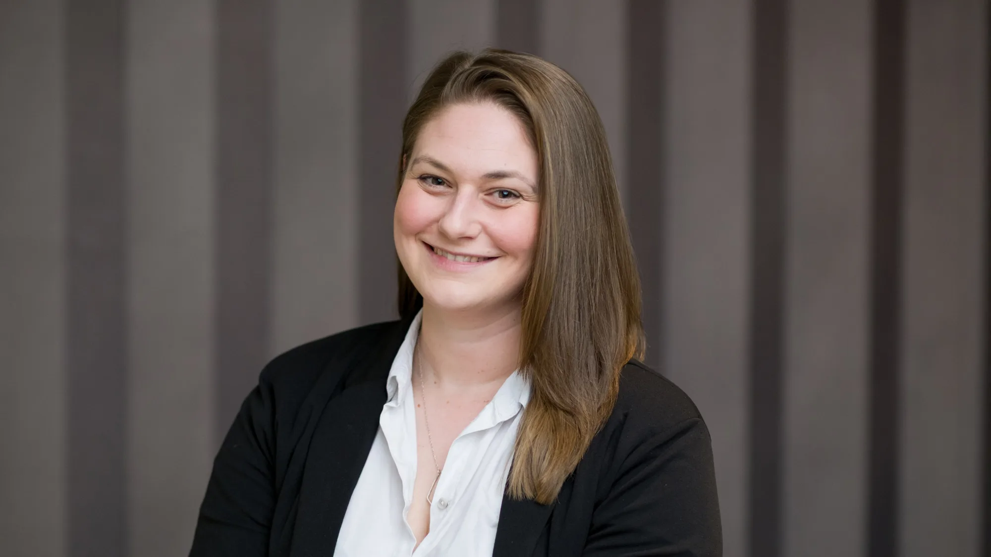 A person with shoulder-length brown hair, wearing a white button-up shirt and black blazer, seated in front of a background with vertical grey stripes.