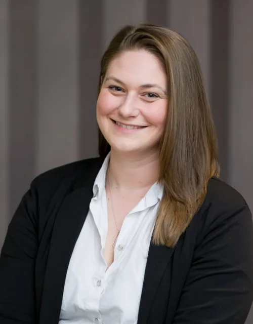 A person with shoulder-length brown hair, wearing a white button-up shirt and black blazer, seated in front of a background with vertical grey stripes.