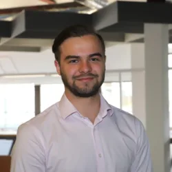A person with short hair and a beard wearing a light pink button-up shirt stands in a modern office with large windows and ceiling light fixtures.