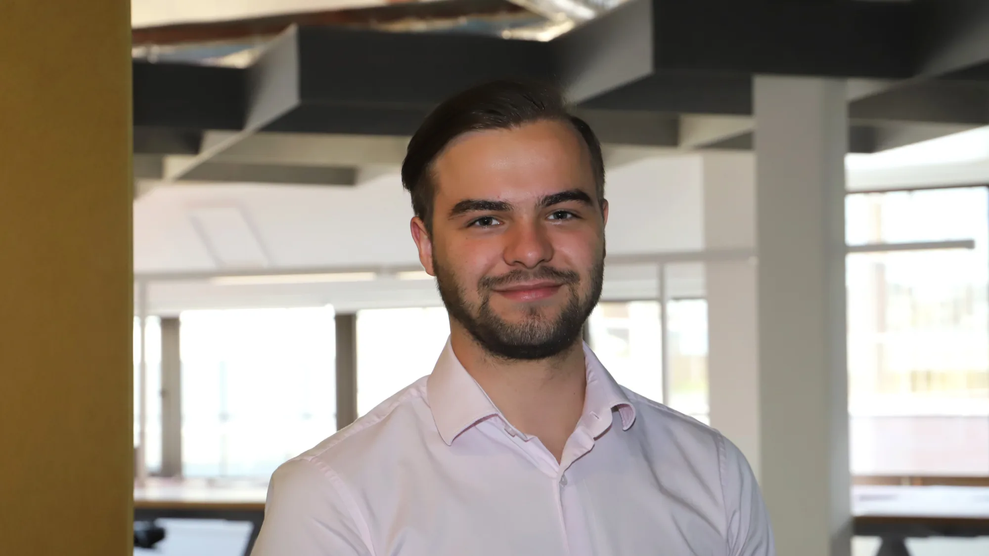 A person with short hair and a beard wearing a light pink button-up shirt stands in a modern office with large windows and ceiling light fixtures.