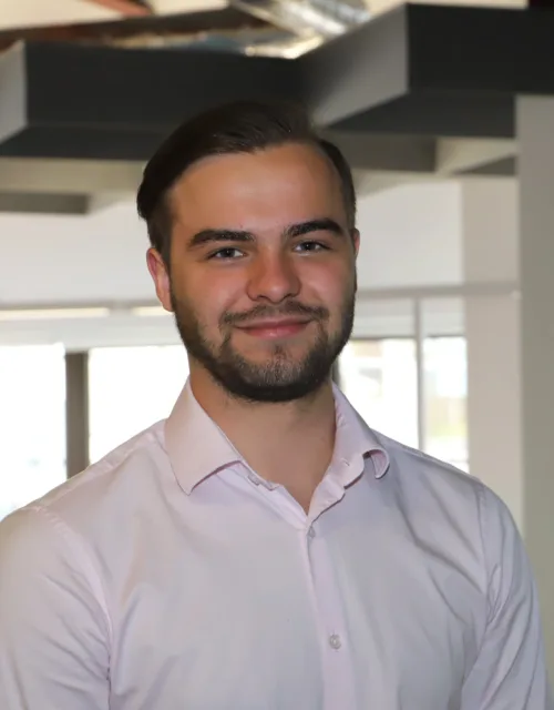 A person with short hair and a beard wearing a light pink button-up shirt stands in a modern office with large windows and ceiling light fixtures.