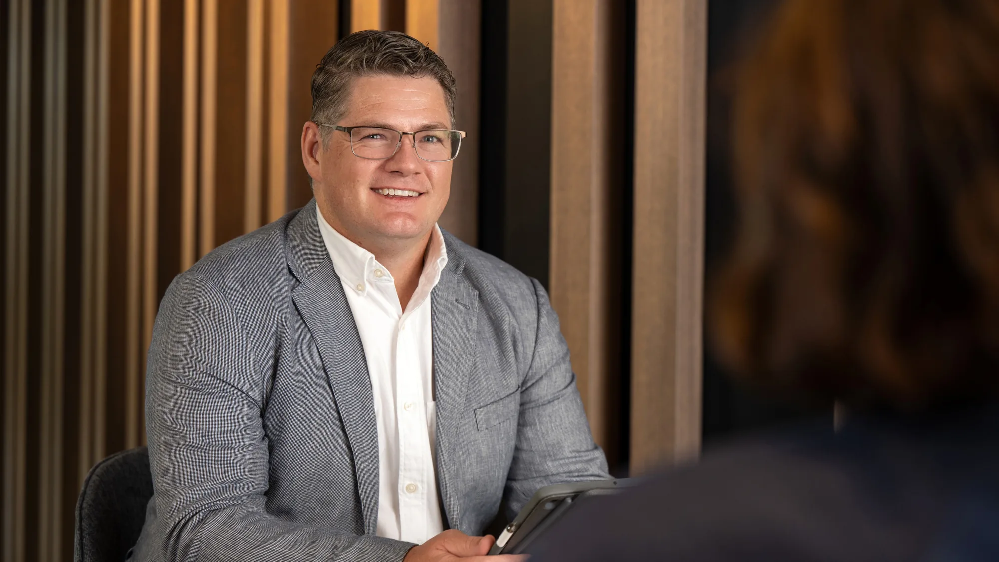 David Collins wearing a white shirt and grey suit jacket sat down talking to someone with their back turned