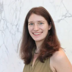 Head-and-shoulders portrait of a person with shoulder-length reddish-brown hair, facing the camera with teeth visible, wearing a sleeveless olive-green top, standing against a light marble-patterned wall.