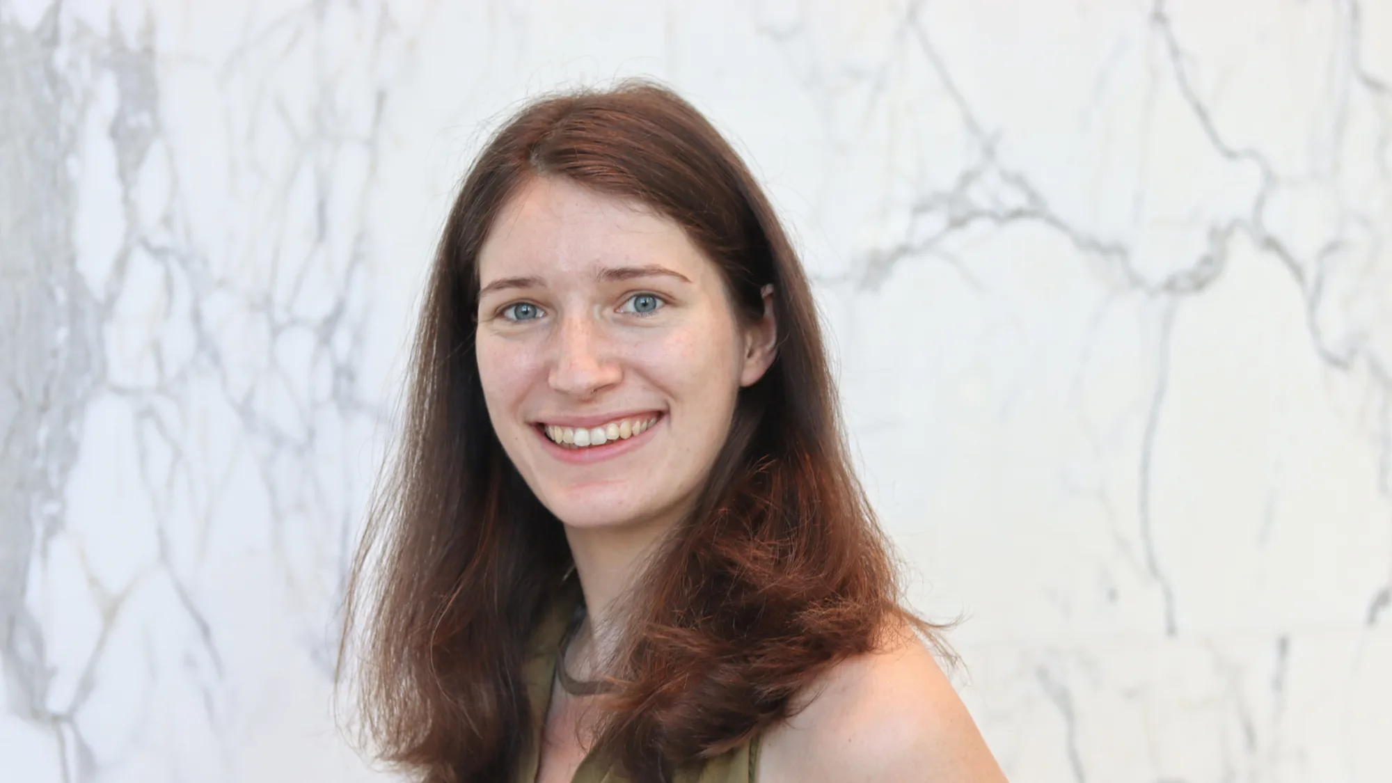Head-and-shoulders portrait of a person with shoulder-length reddish-brown hair, facing the camera with teeth visible, wearing a sleeveless olive-green top, standing against a light marble-patterned wall.