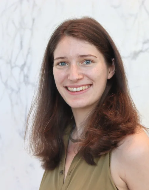 Head-and-shoulders portrait of a person with shoulder-length reddish-brown hair, facing the camera with teeth visible, wearing a sleeveless olive-green top, standing against a light marble-patterned wall.