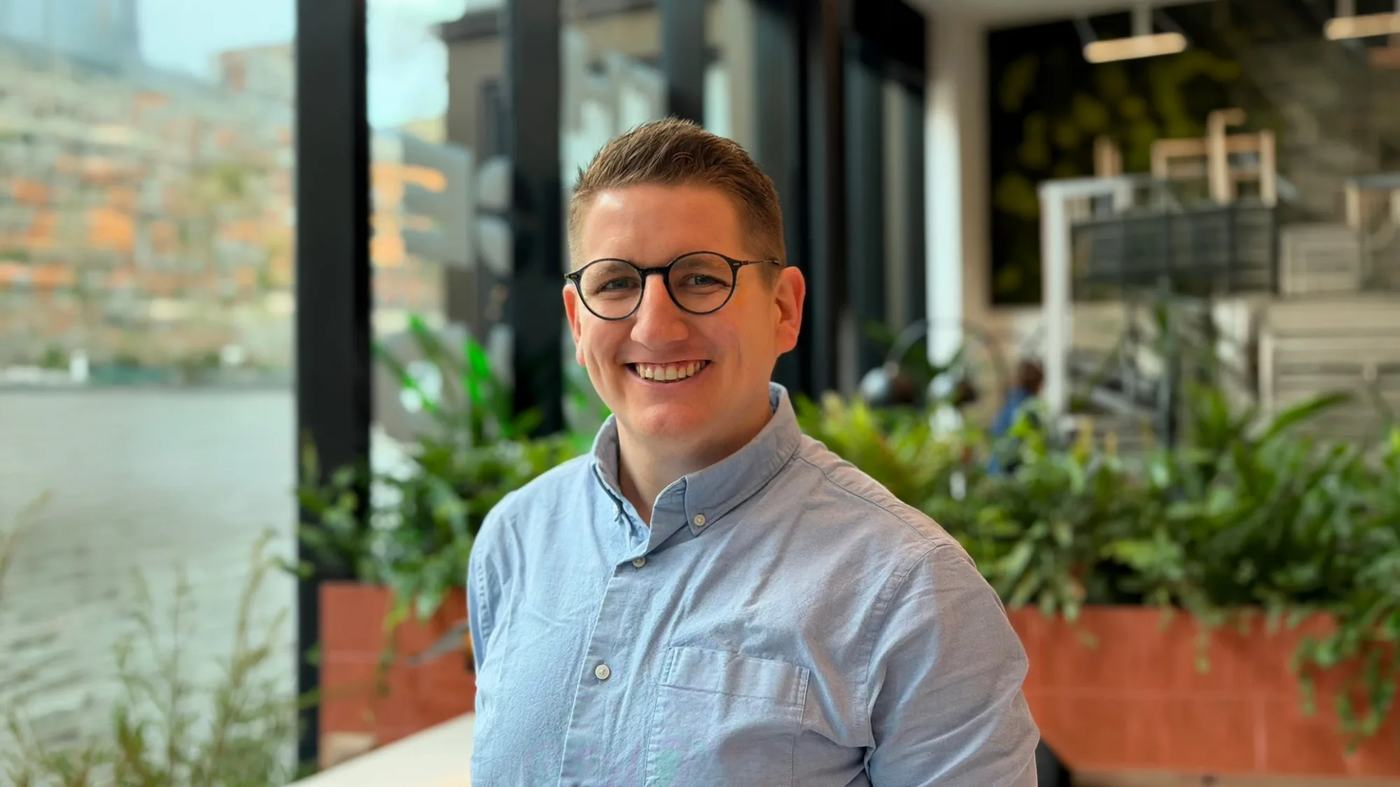 A person wearing round glasses and a light blue button-up shirt is shown from the chest up indoors, standing near large windows with black frames, with potted plants and a blurred cityscape visible in the background.