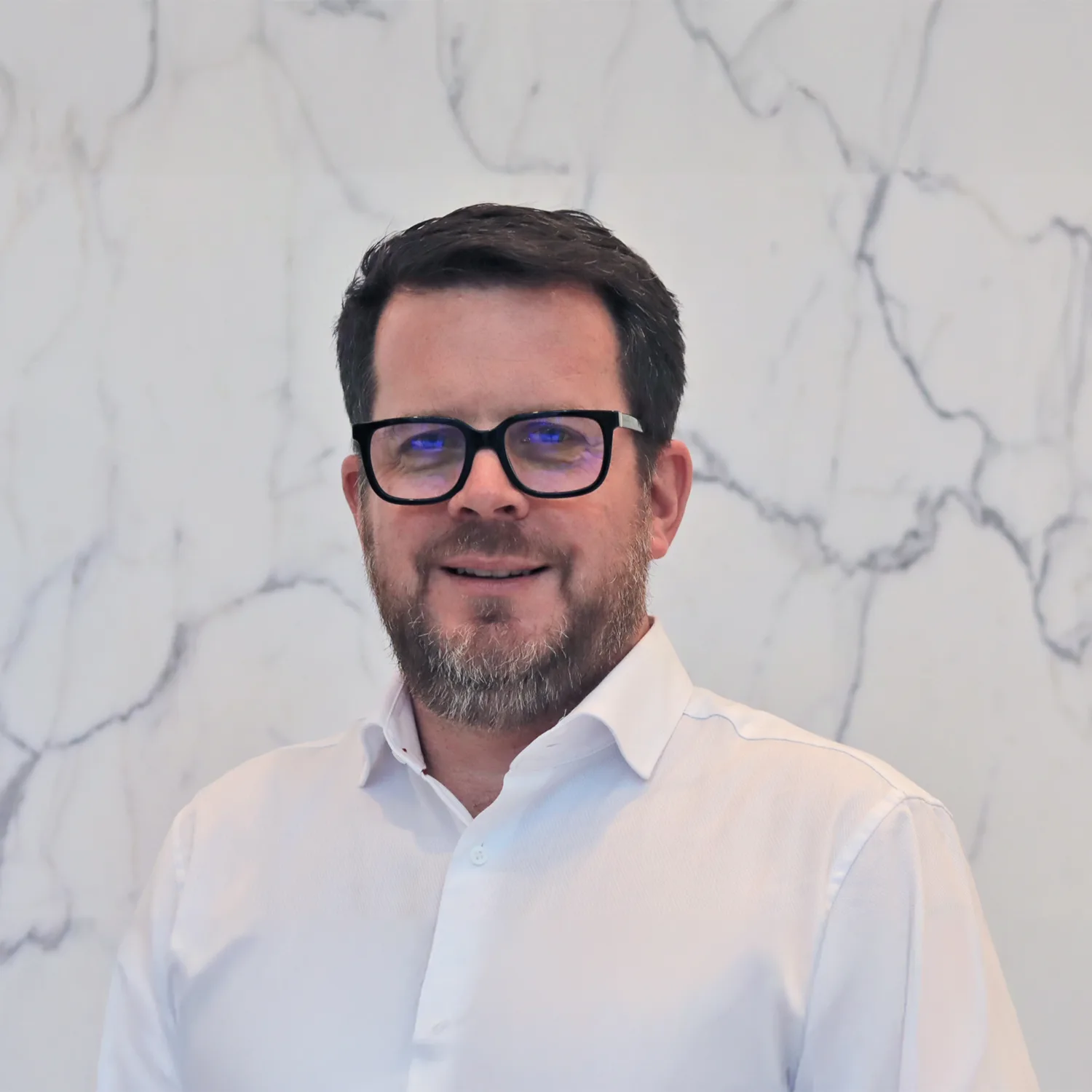Head and sholders shot of Paul, wearing a white shirt and glasses against a white marble background.