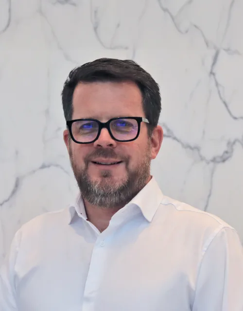 Head and sholders shot of Paul, wearing a white shirt and glasses against a white marble background.