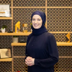 Bysshe Wallace standing indoors in front of a decorative wall with circular patterns and wooden shelves displaying framed certificates, small plants, mugs, and geometric objects; red potted plants on the floor.