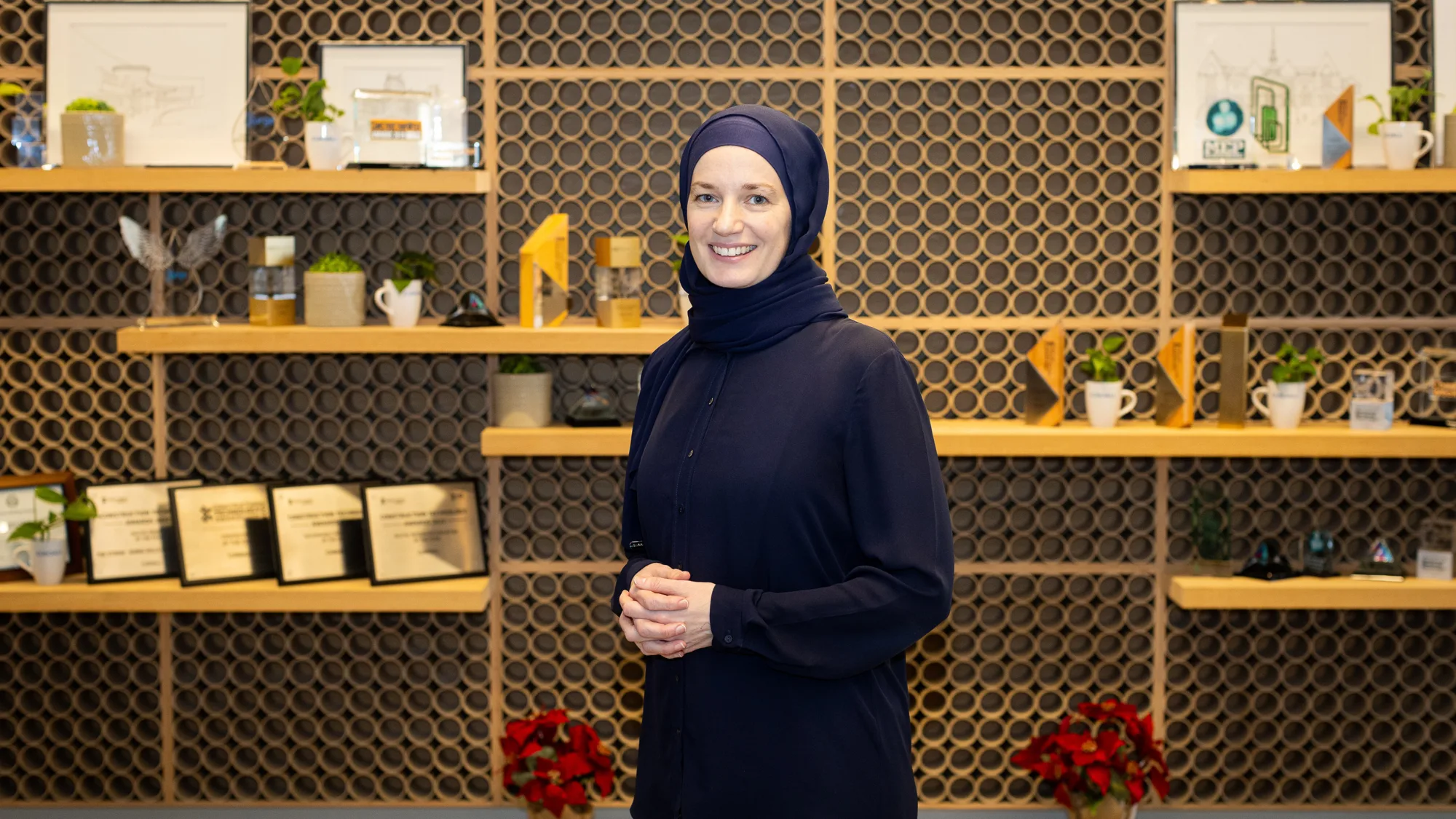 Bysshe Wallace standing indoors in front of a decorative wall with circular patterns and wooden shelves displaying framed certificates, small plants, mugs, and geometric objects; red potted plants on the floor.