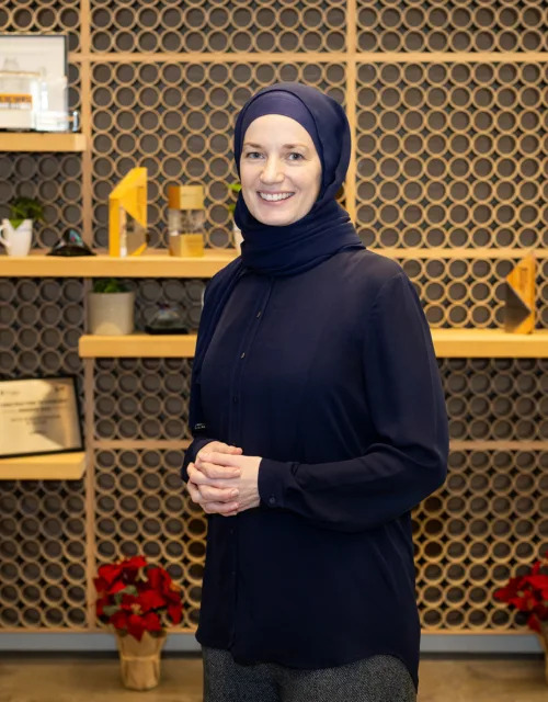 Bysshe Wallace standing indoors in front of a decorative wall with circular patterns and wooden shelves displaying framed certificates, small plants, mugs, and geometric objects; red potted plants on the floor.
