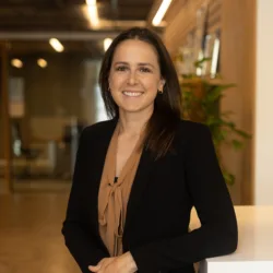 Annabelle standing indoors wearing a black blazer over a brown blouse. The background shows an office with glass walls, ceiling lights, and a plant on the right side.